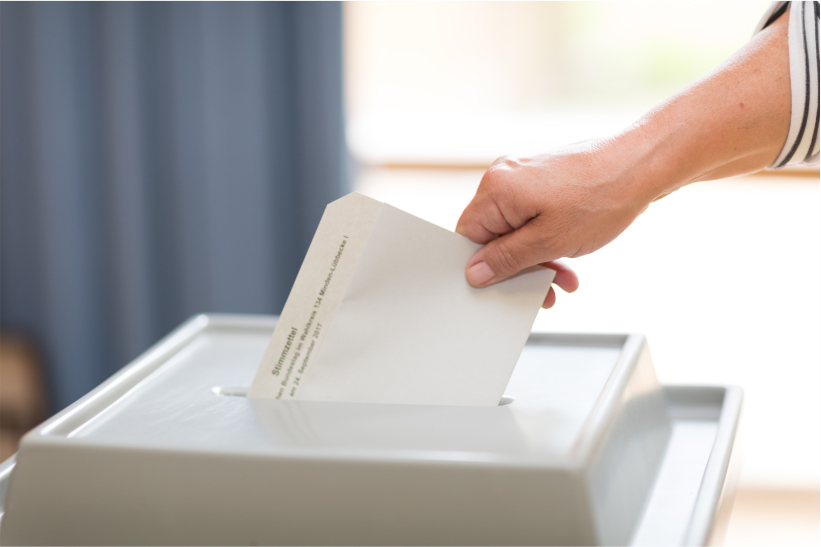 Ein Wahlzettel wird in eine Urne geworfen als Hinweis auf das Thema Wahlen